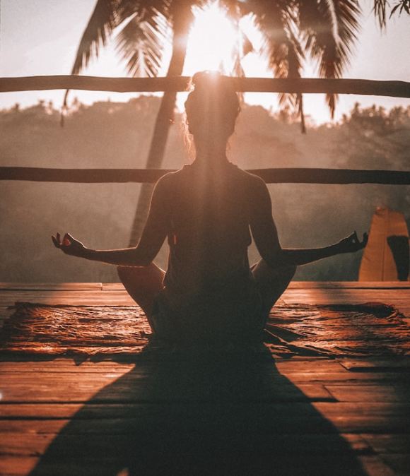 Femme en posture de yoga sur un balcon en bois, devant le soleil couchant.