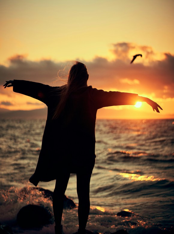 Femme en équilibre, face au soleil couchant sur la mer. Cette femme sent libérée de ses tensions. Ses cheveux et sa robe volent légèrement au vent, une mouette passe dans le ciel oranger.